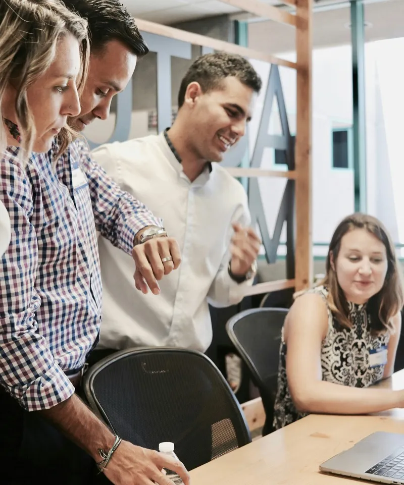 Four colleagues gathered around a laptop at a shared desk, smiling and talking.